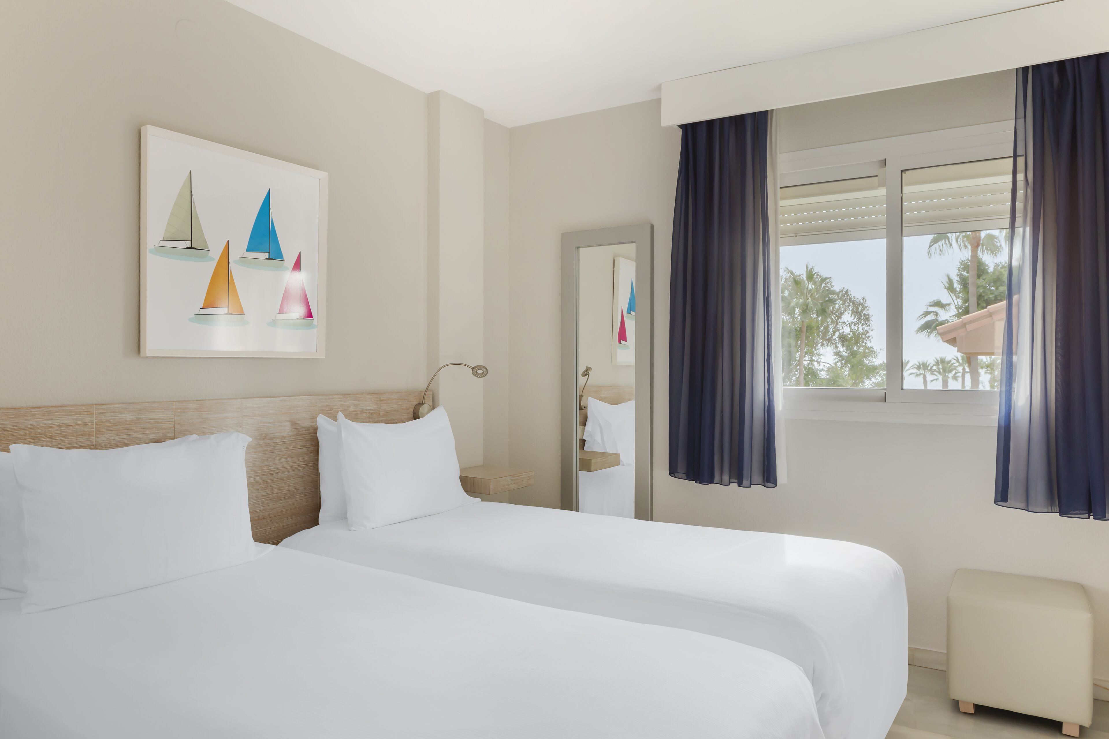 Una habitación de hotel con dos camas, una ventana con cortinas, un cuadro de veleros y un taburete pequeño.