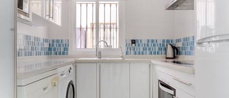 A modern kitchen with white cabinets, a built-in washing machine, a stainless steel sink, and a window with a view of a balcony.