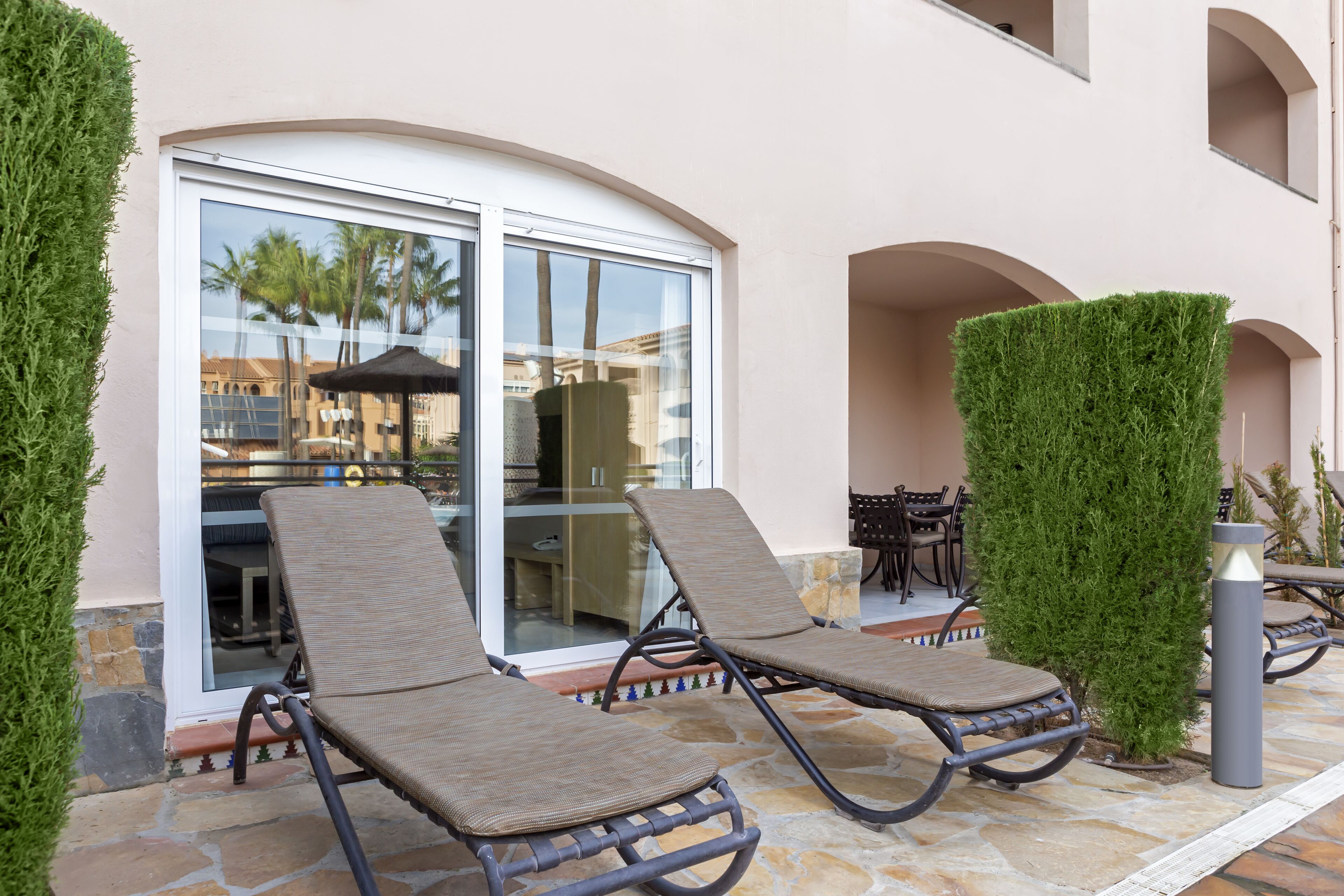 A patio with two lounge chairs, a glass door leading to an indoor area, and a stone floor.