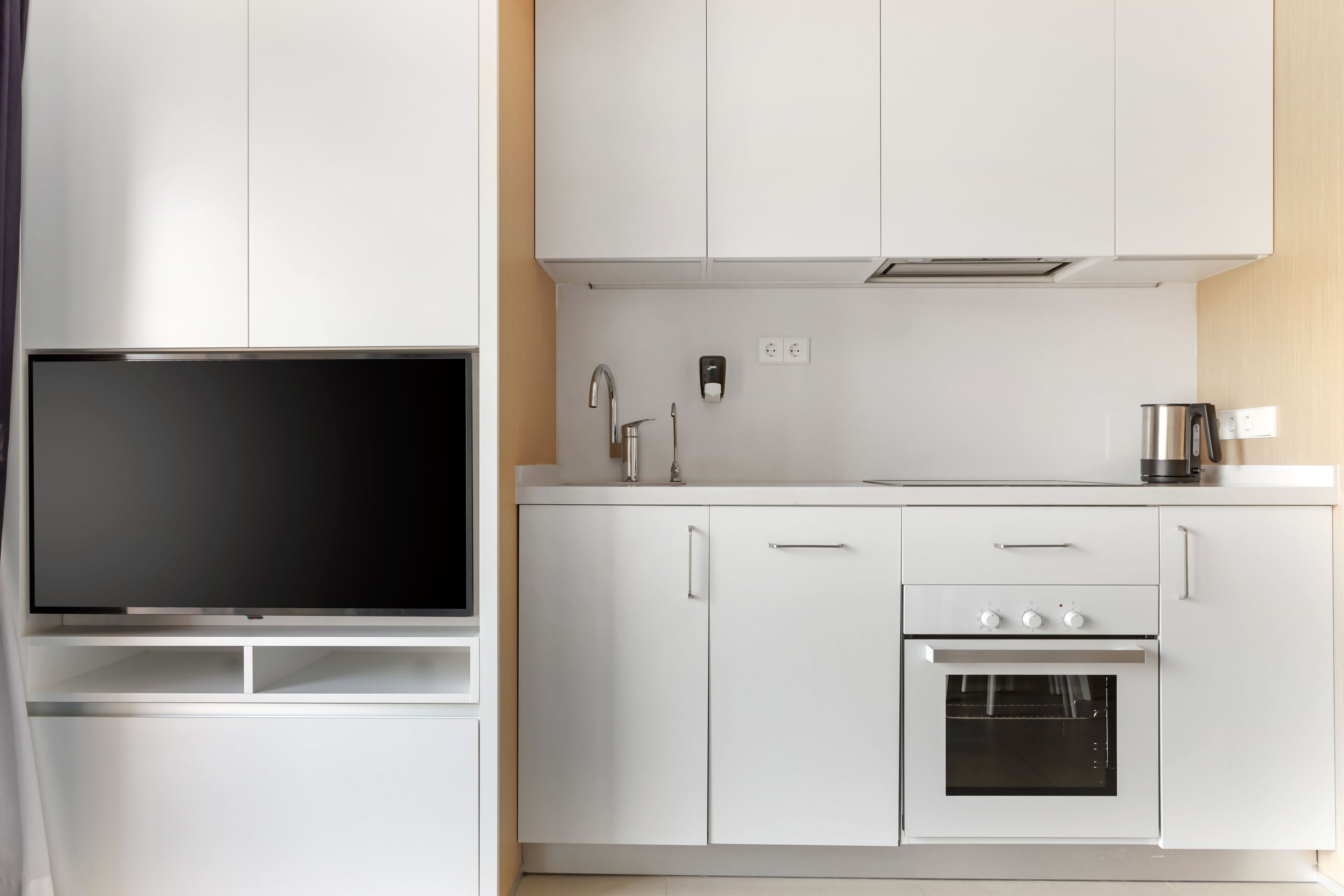 A modern kitchen with white cabinets, a built-in oven, a sink, and a flat-screen TV.
