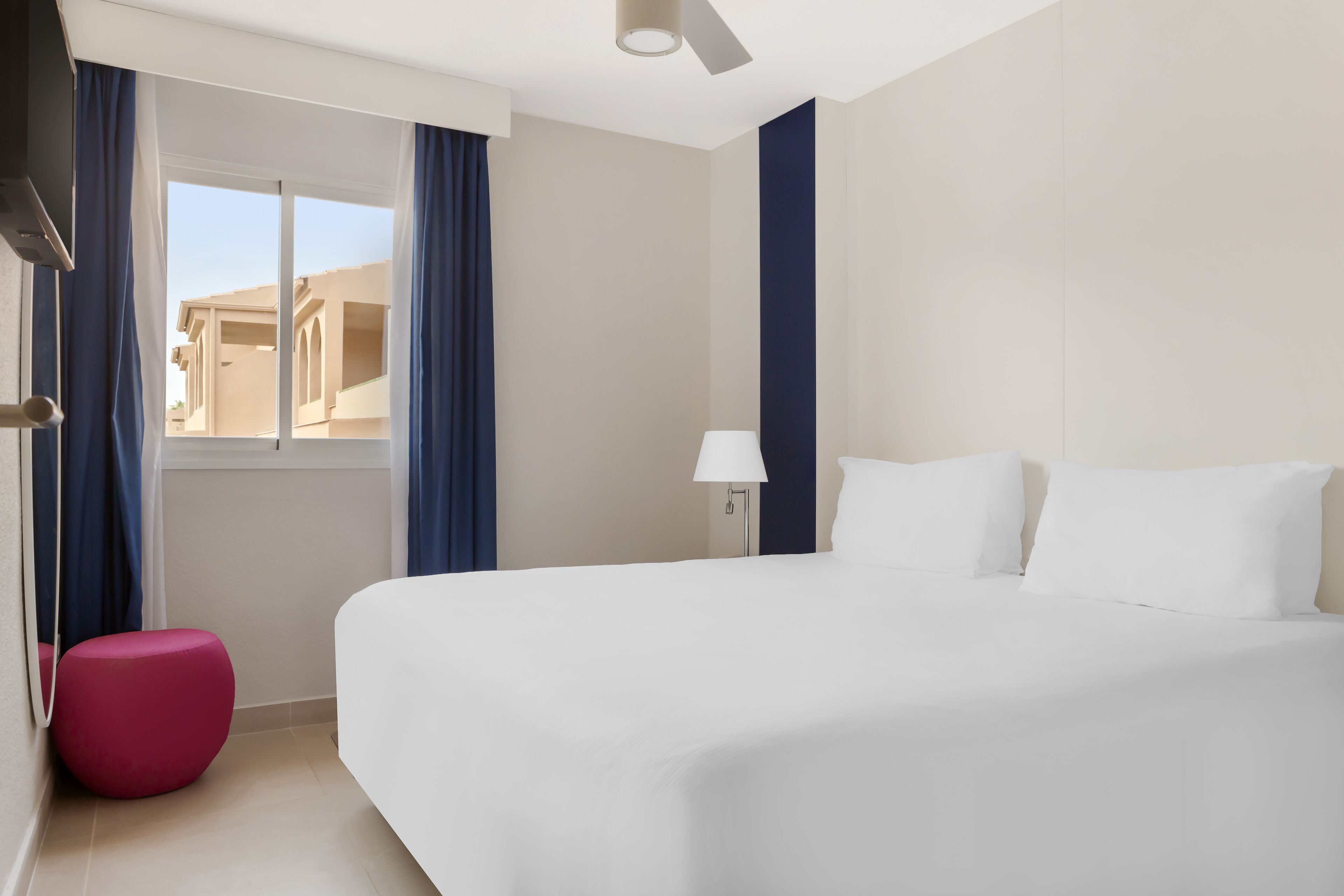 A hotel room with a large bed, a small pink ottoman, a window with curtains, and a lamp.