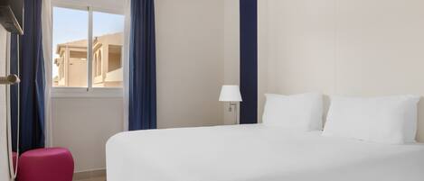 A hotel room with a large bed, a small pink ottoman, a window with curtains, and a lamp.
