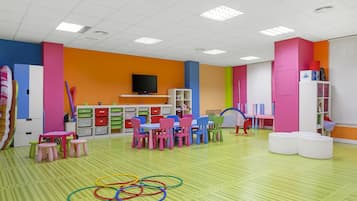 A brightly colored classroom with a green-striped floor, multiple tables and chairs, and educational materials on shelves.