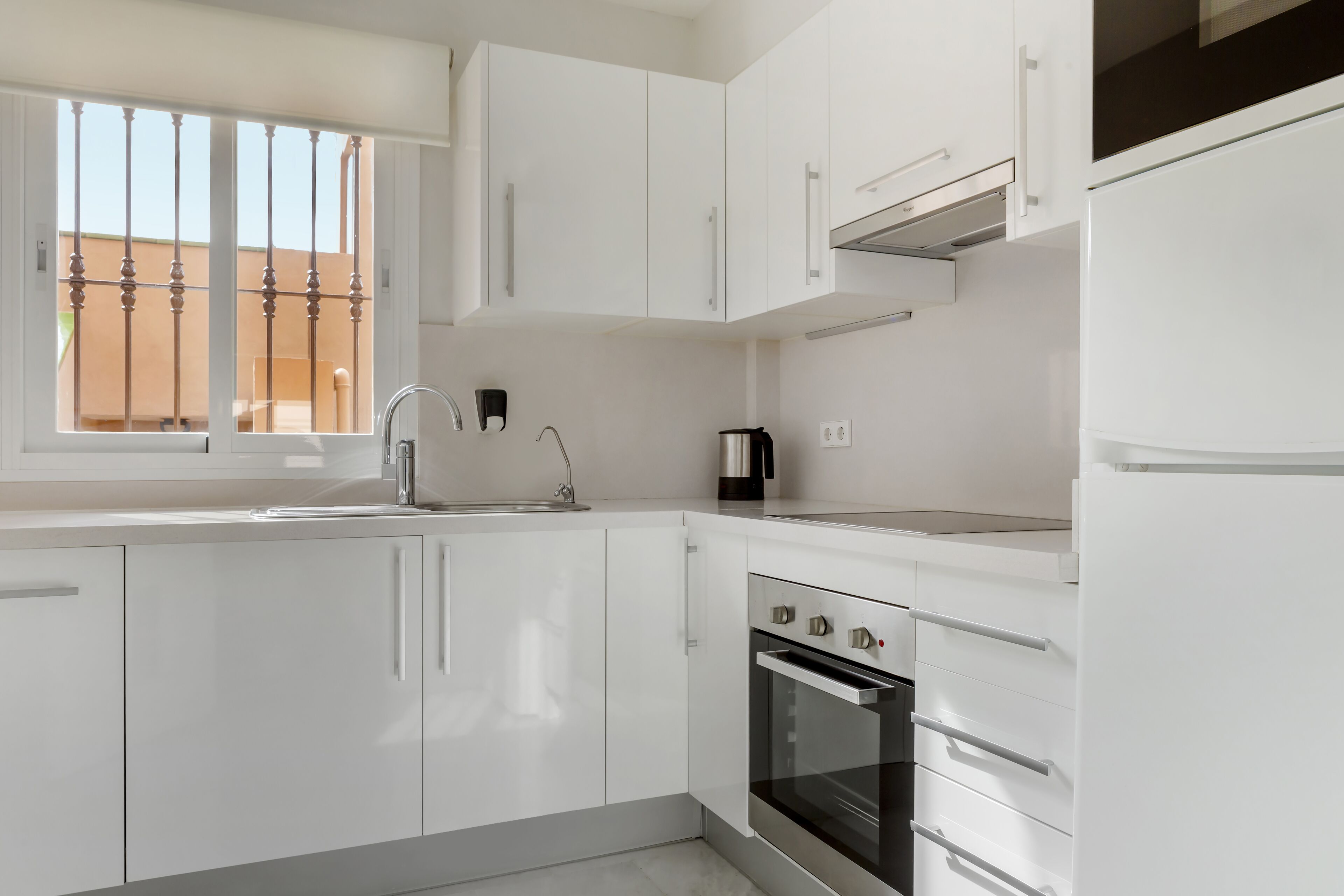 A modern kitchen with white cabinets, a stainless steel oven, and a window with a metal grill.