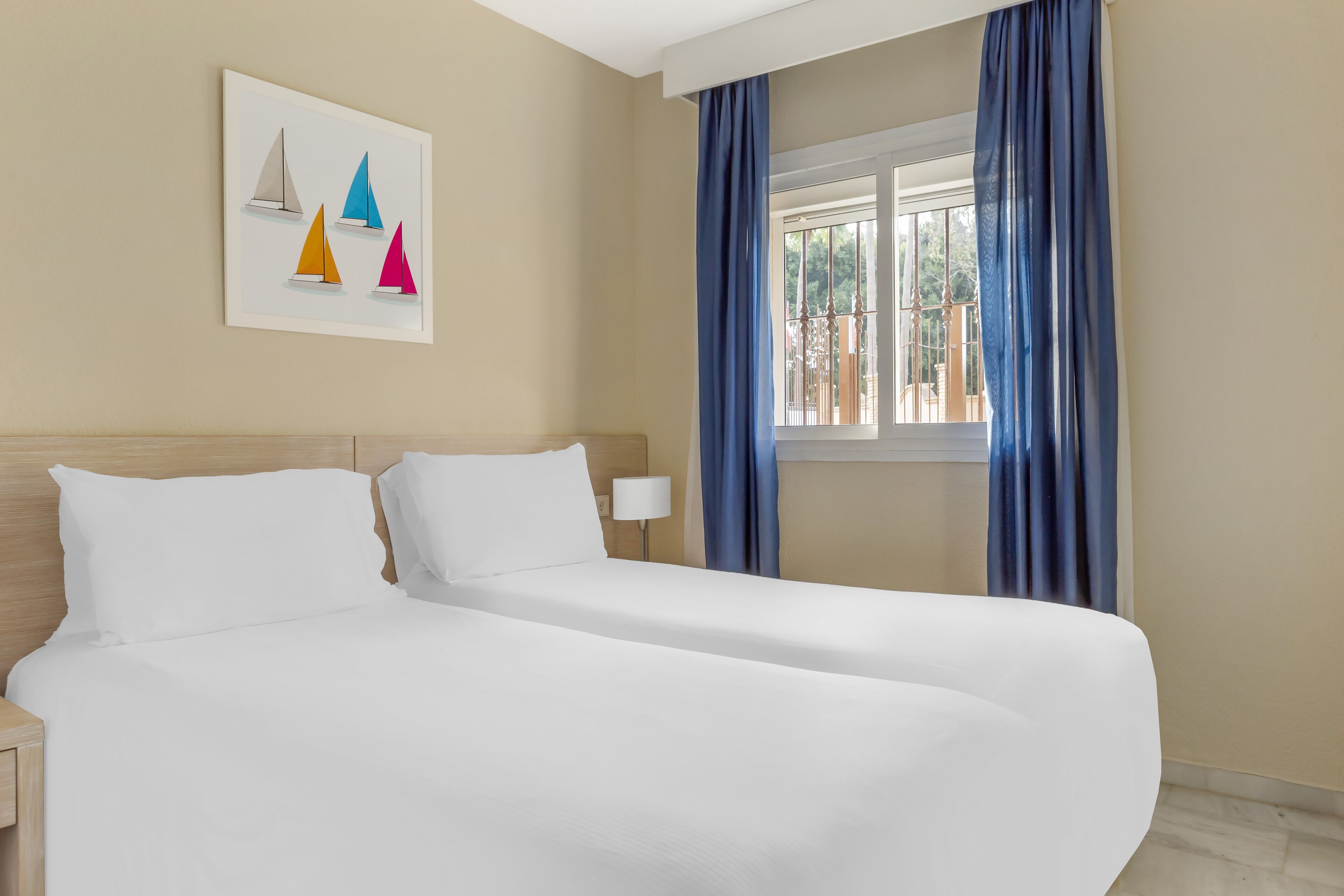 A hotel room with a double bed, a nightstand, a lamp, a window with blue curtains, and a wall art piece of sailboats.