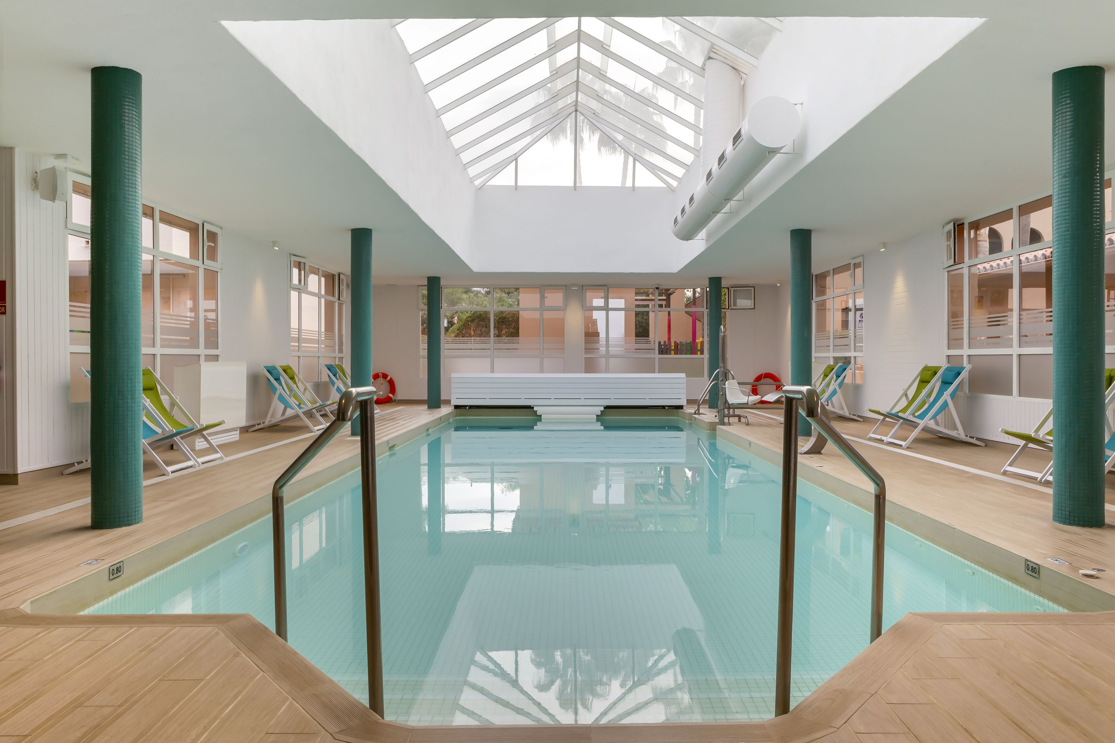 Una piscina interior con techo de vidrio transparente, rodeada de sillas de descanso y suelo de baldosas.