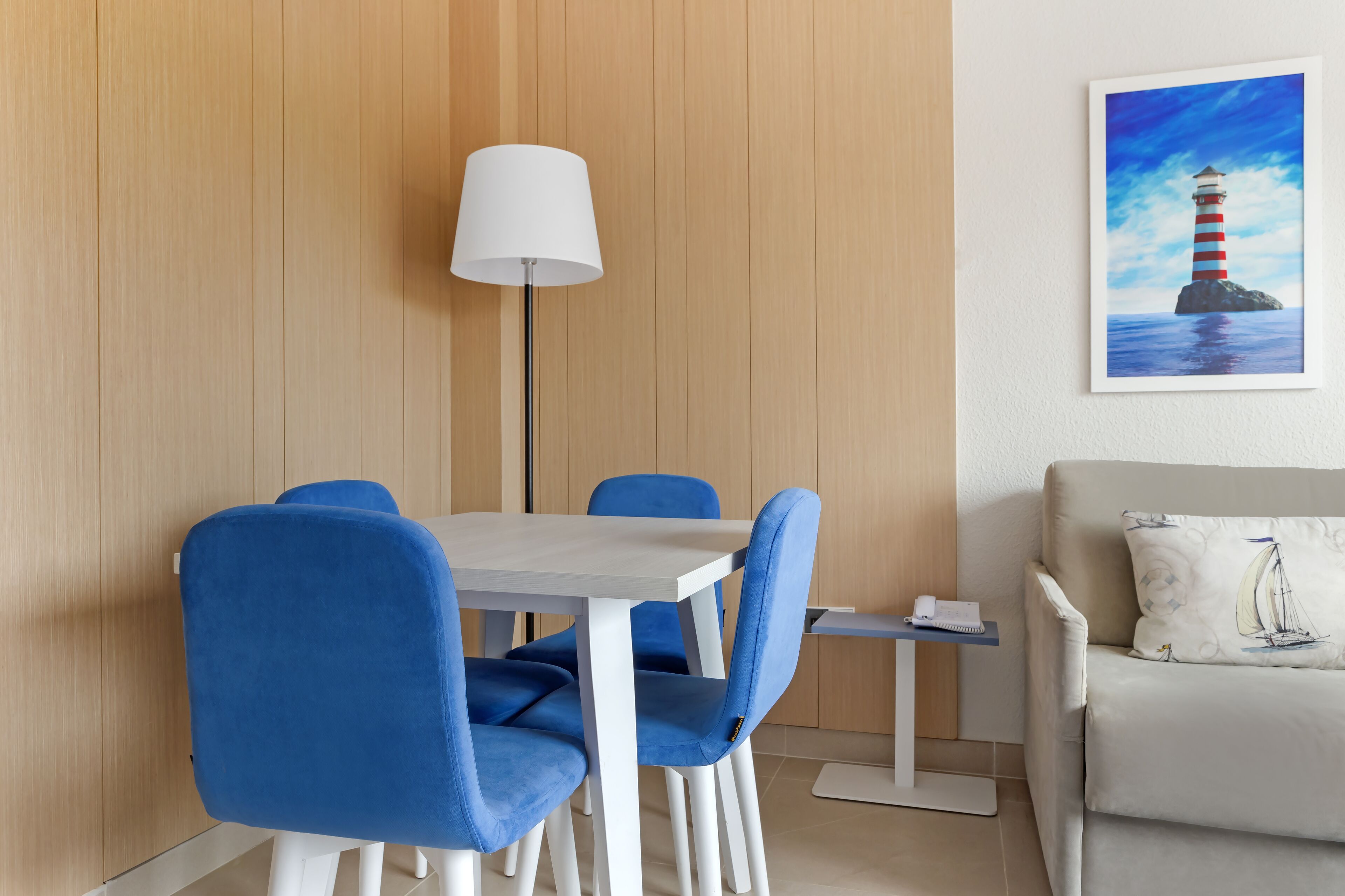 A dining area with a white table and blue chairs, a floor lamp, and a painting of a lighthouse on the wall.