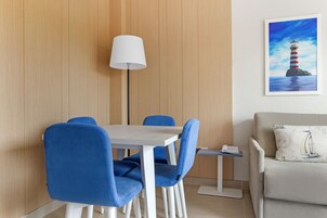 A dining area with a white table and blue chairs, a floor lamp, and a painting of a lighthouse on the wall.