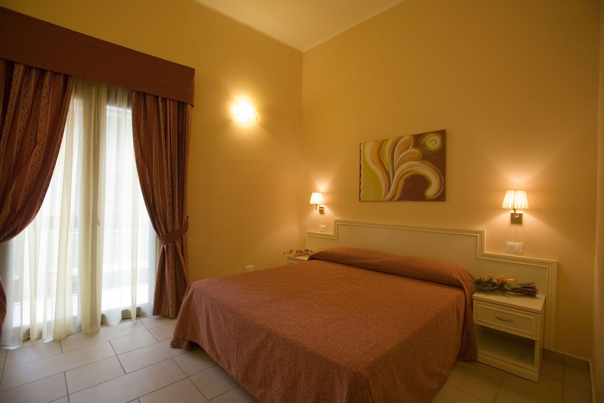 Ein Hotelzimmer mit einem großen Bett, zwei Nachttischen, einem Wandbild und einem Fenster mit Vorhängen.