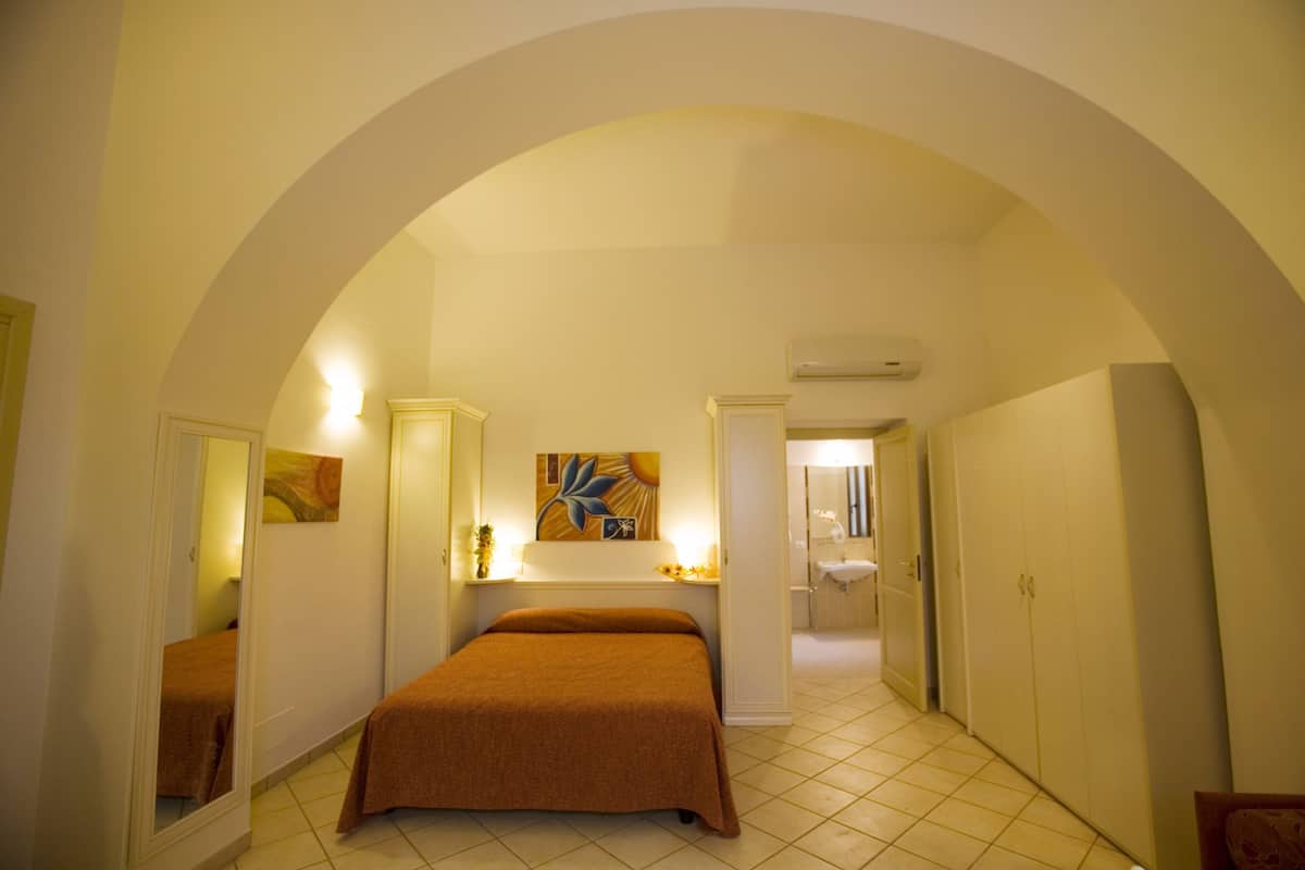 Ein Schlafzimmer mit Bett, Spiegel, Gemälde und angrenzendem Badezimmer, durchbrochen von einem Bogen.