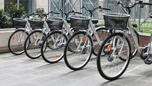 Una hilera de bicicletas blancas con cestas negras, estacionadas frente a un edificio con valla metálica.