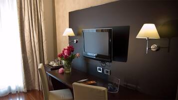 Habitación de hotel con televisor de pantalla plana montado en la pared, escritorio con un jarrón de flores, una silla y dos lámparas de pared.