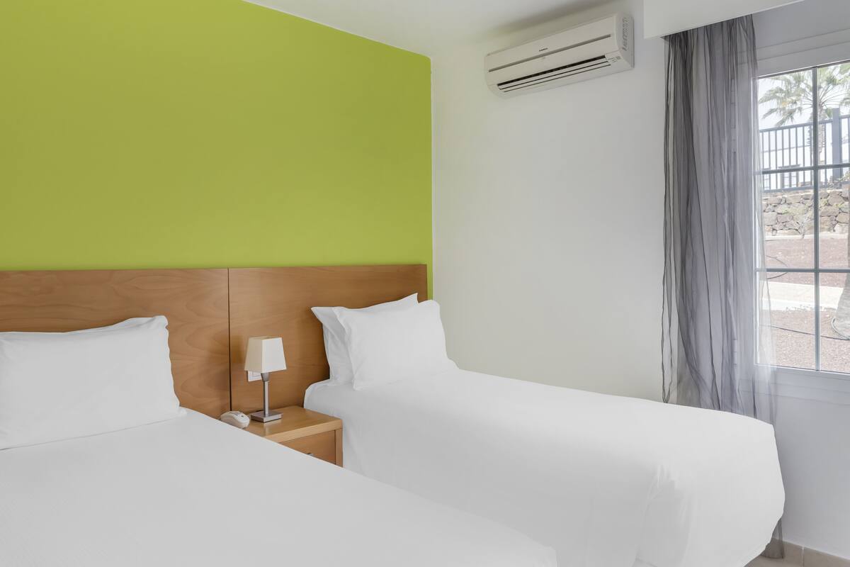 A hotel room with two beds, a wooden headboard, a nightstand with a lamp, and an air conditioning unit.