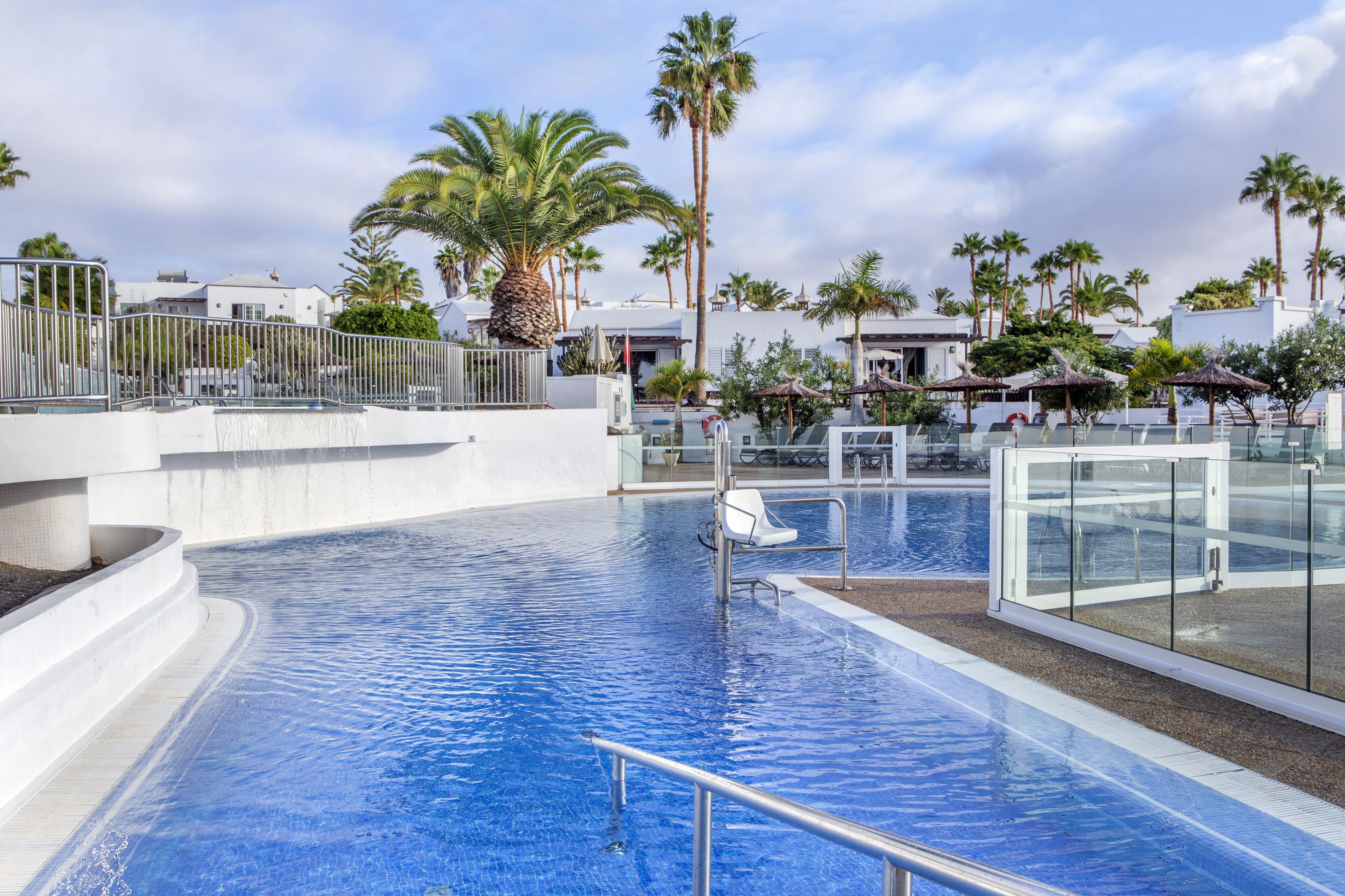 A swimming pool with a glass barrier, palm trees, and modern buildings in the background.