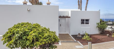 A modern building with a white exterior, a small window, and a door. There are palm trees and a clear sky in the background.