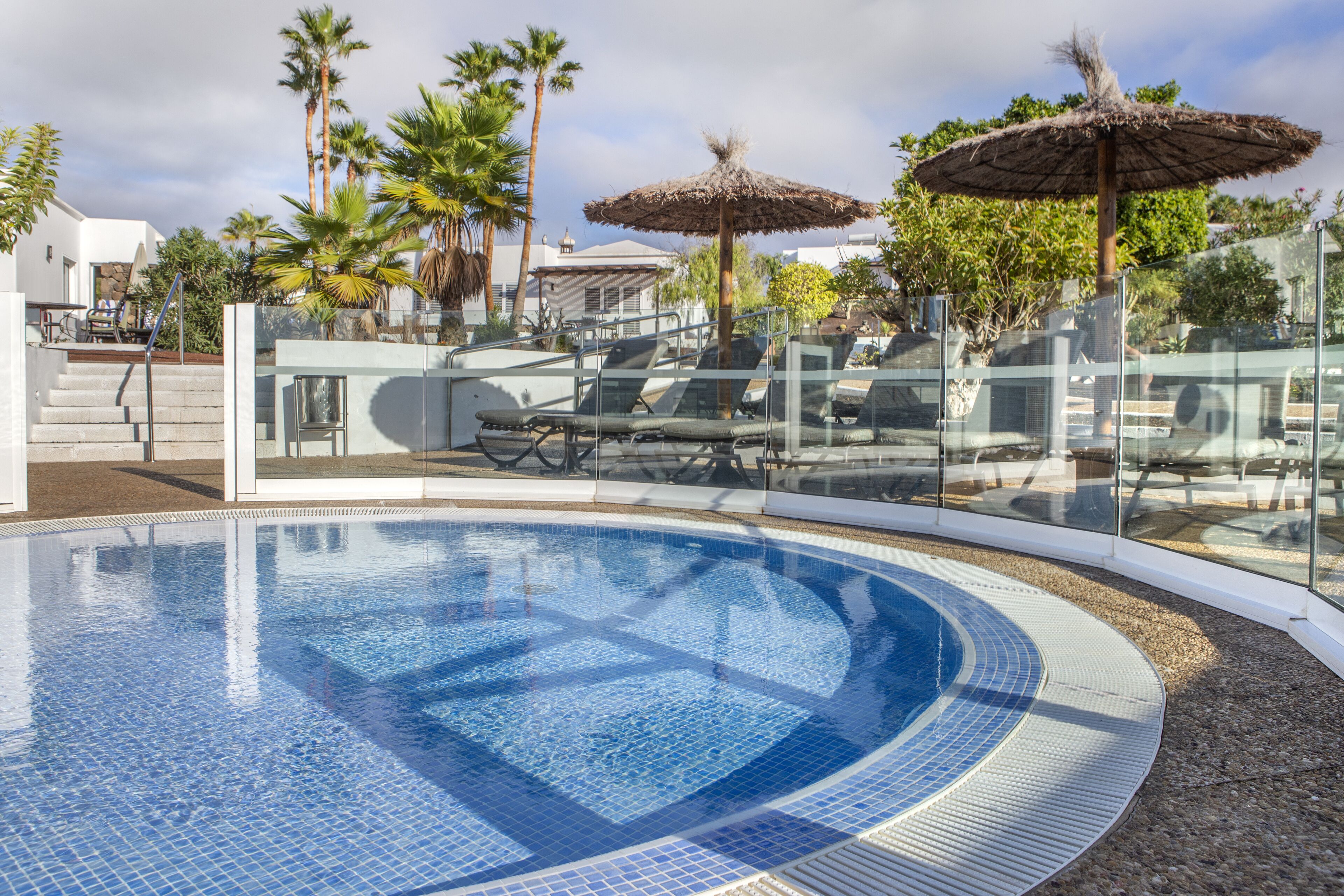 Una piscina con sillas de descanso y sombrillas, rodeada de palmeras y una cerca de vidrio.