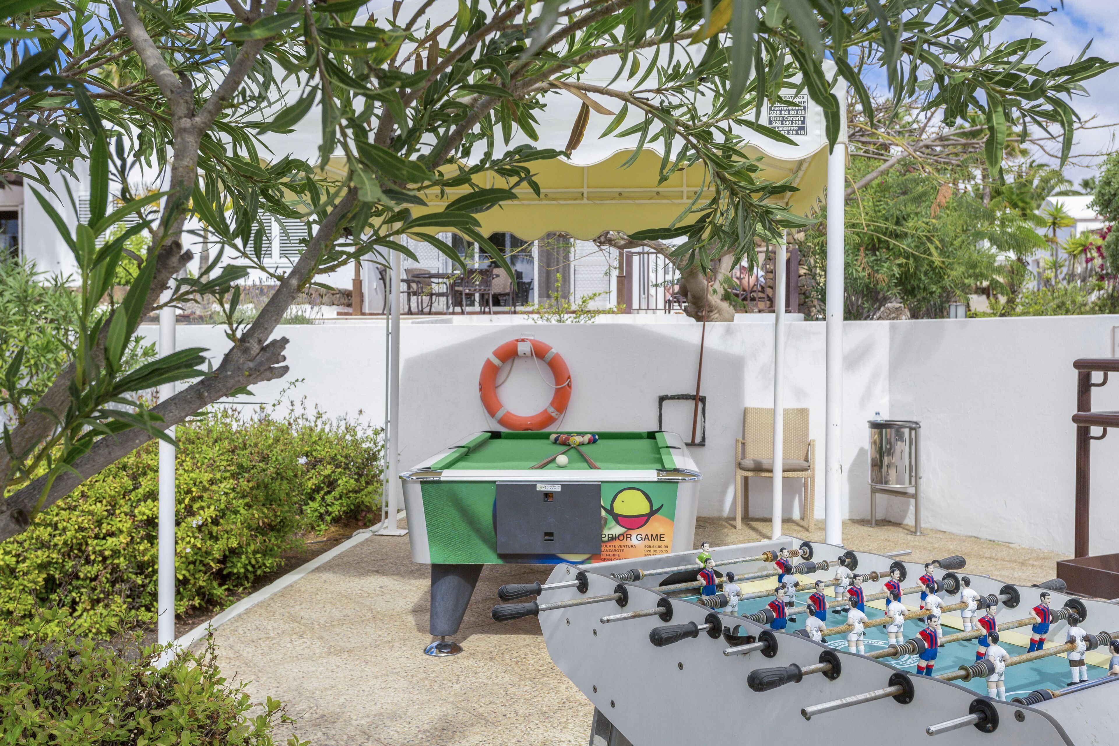 An outdoor foosball table with a green table and a white backboard, surrounded by greenery and a white wall.