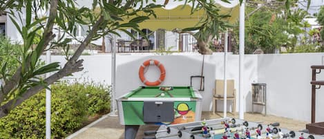 An outdoor foosball table with a green table and a white backboard, surrounded by greenery and a white wall.