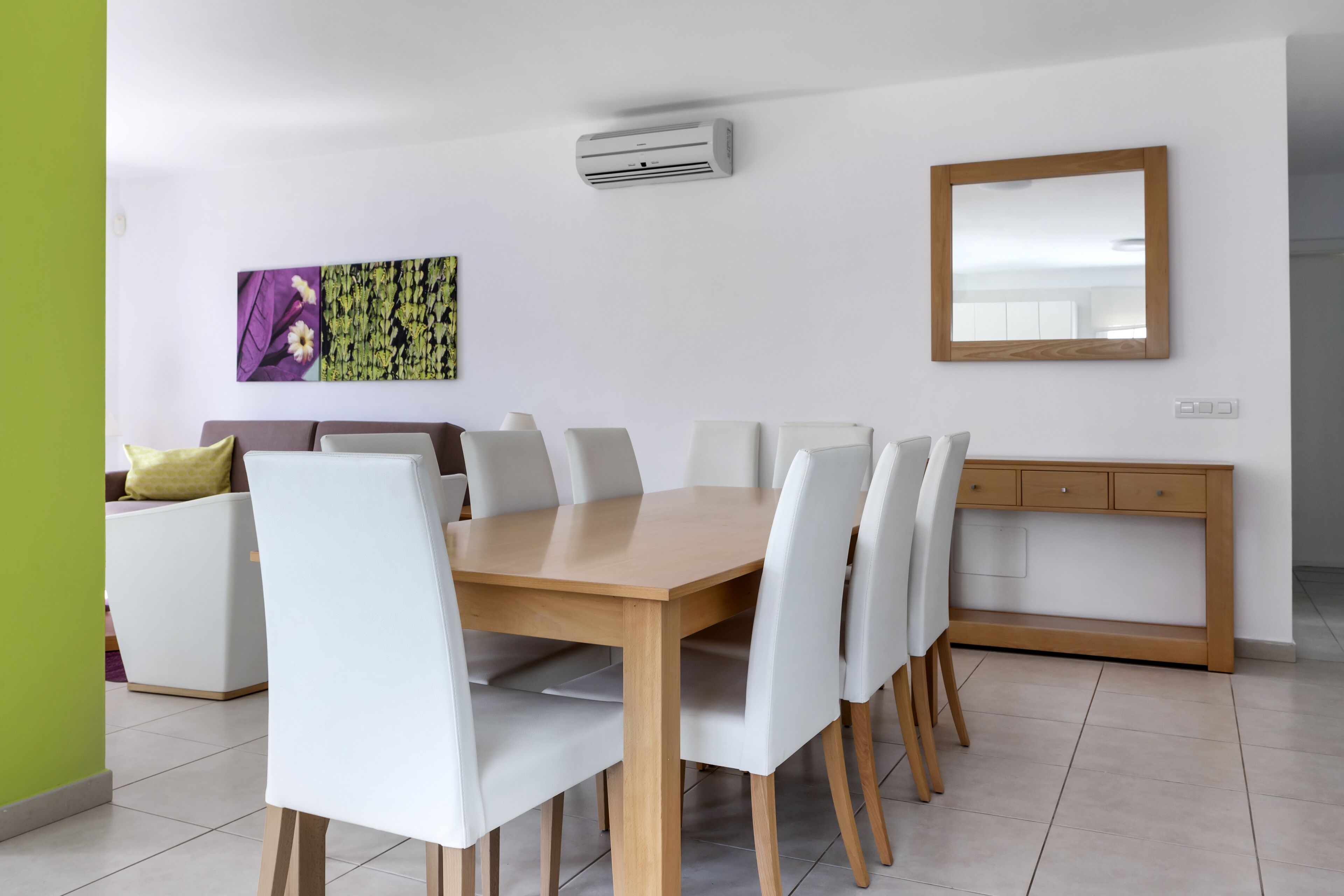 A dining area with a wooden table and white chairs, a sofa, a cabinet, and a wall-mounted air conditioner.