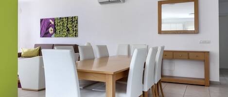 A dining area with a wooden table and white chairs, a sofa, a cabinet, and a wall-mounted air conditioner.