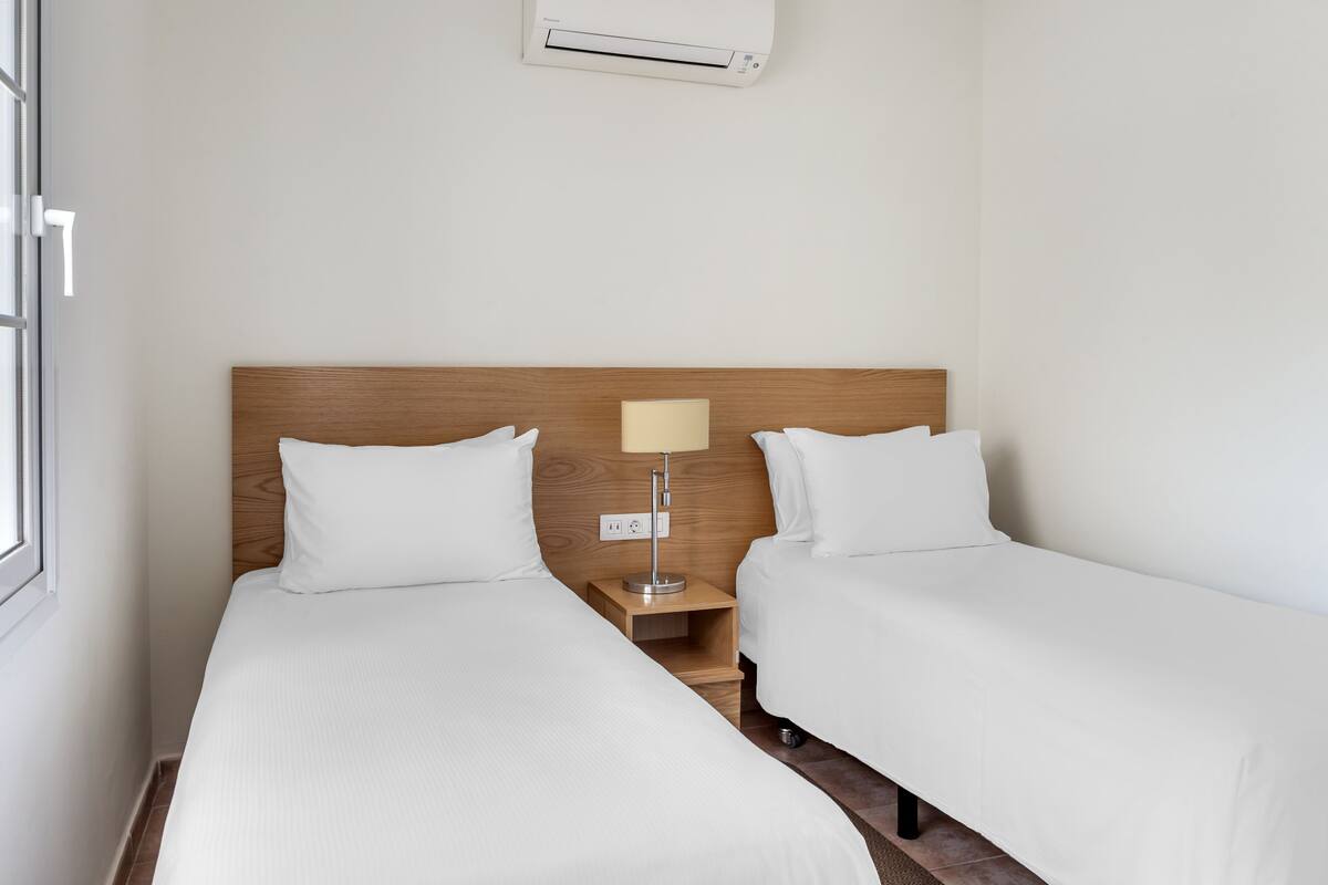 A hotel room with two single beds, a wooden headboard, a bedside table with a lamp, and a wall-mounted air conditioner.