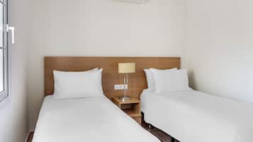 A hotel room with two single beds, a wooden headboard, a bedside table with a lamp, and a wall-mounted air conditioner.