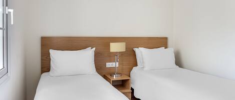 A hotel room with two single beds, a wooden headboard, a bedside table with a lamp, and a wall-mounted air conditioner.