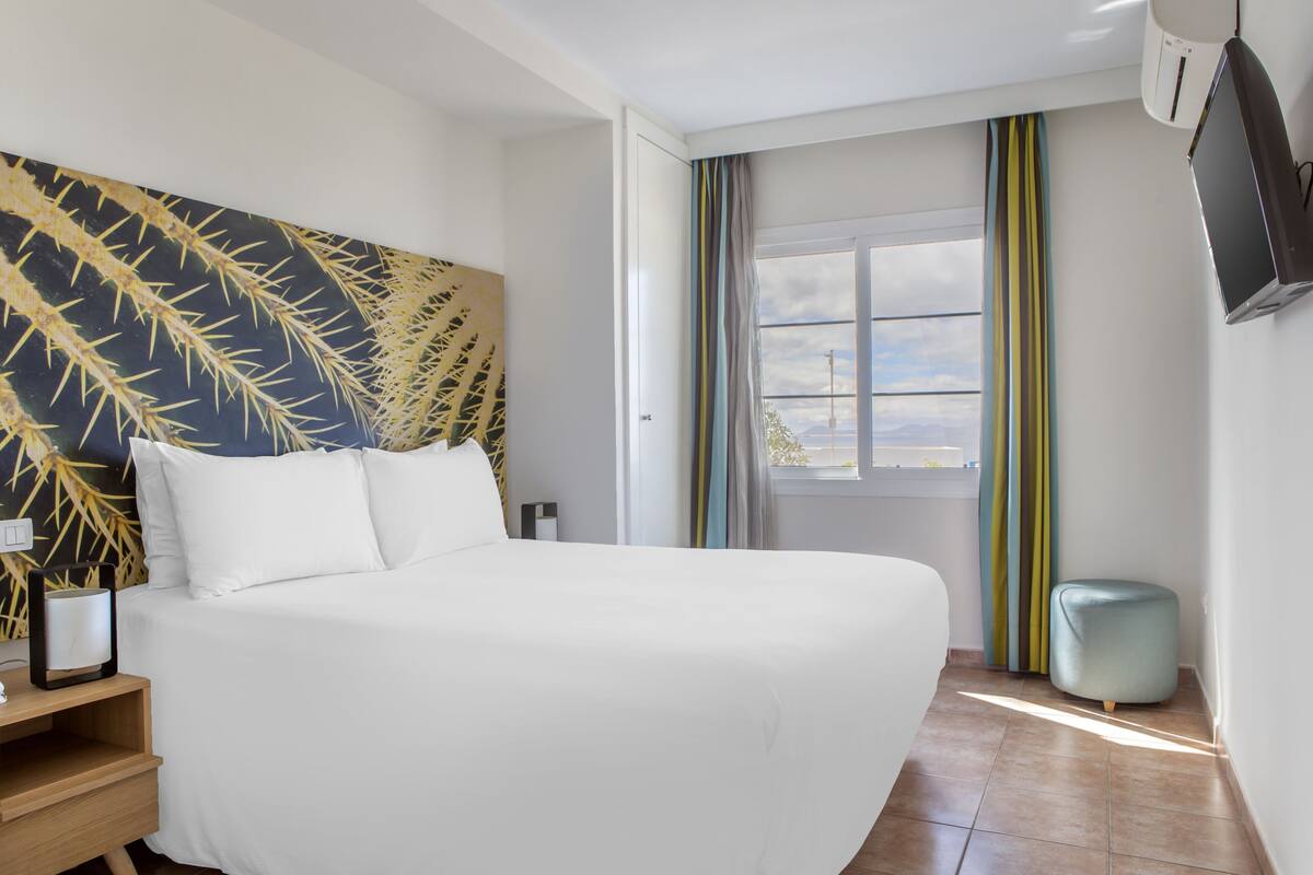 A hotel room with a large bed, a bedside table, a wall-mounted TV, and a window with curtains.