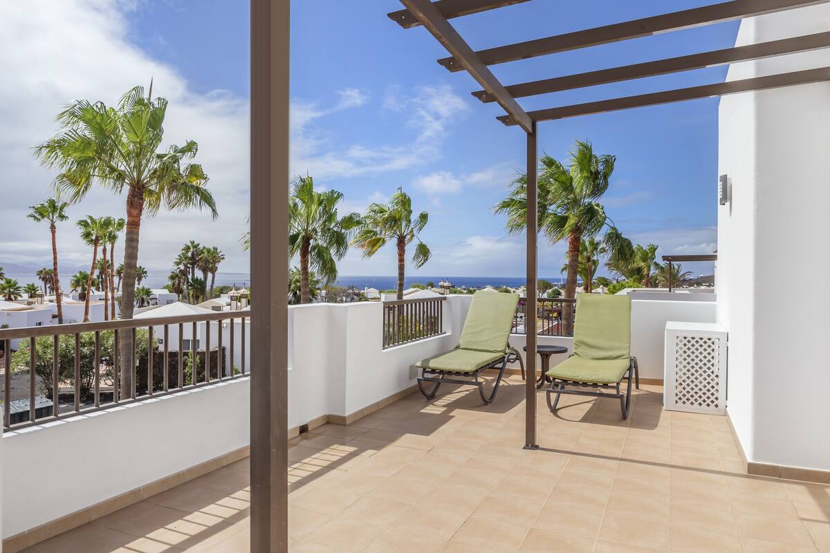 A balcony with two green cushioned chairs, a white wall, and a view of palm trees and the sea.