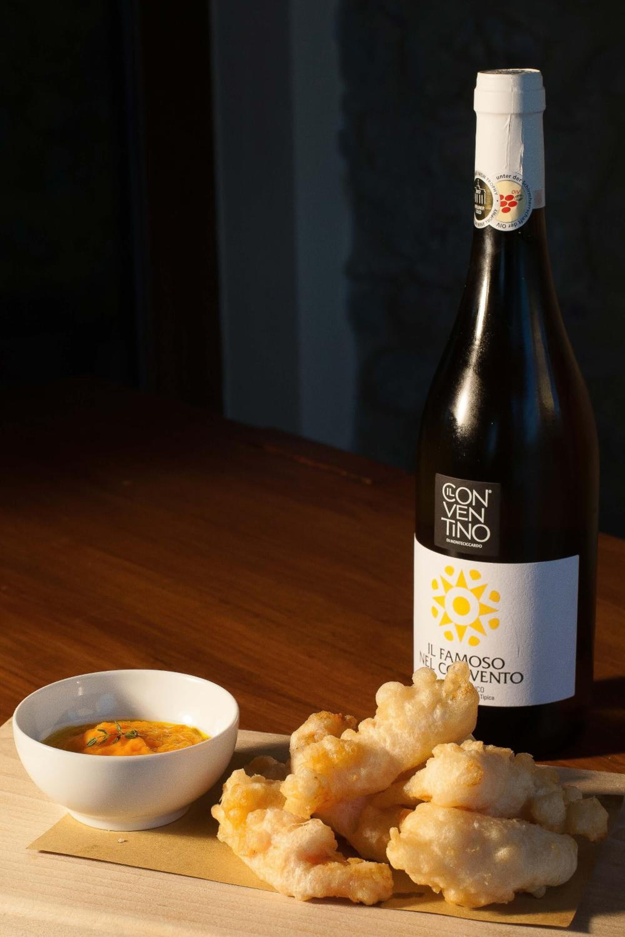 A bottle of Convento Il Famoso wine, a bowl of soup, and breadsticks on a wooden table.