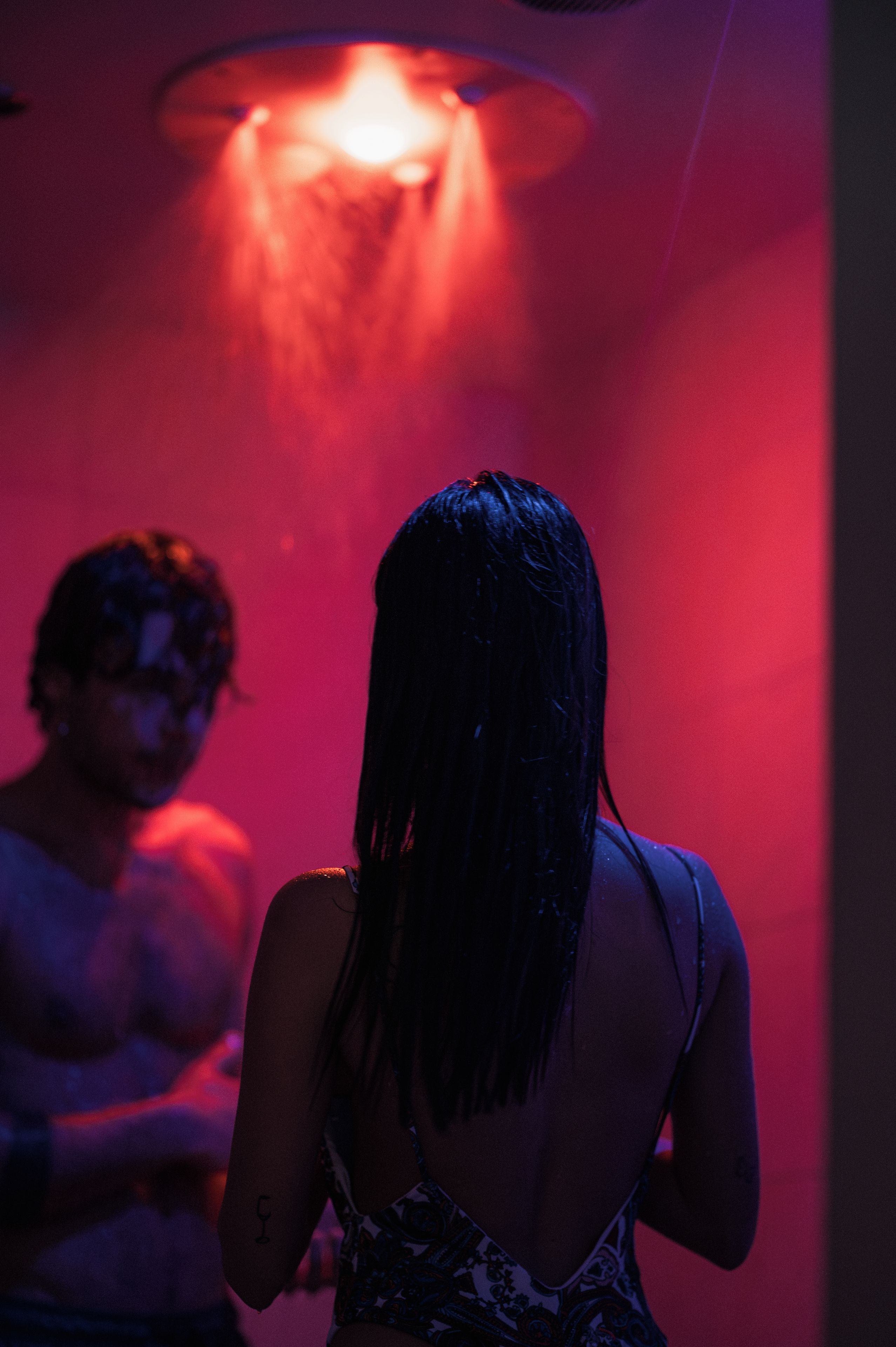 A person with long hair facing away from the camera, standing in a dimly lit room with red lighting.
