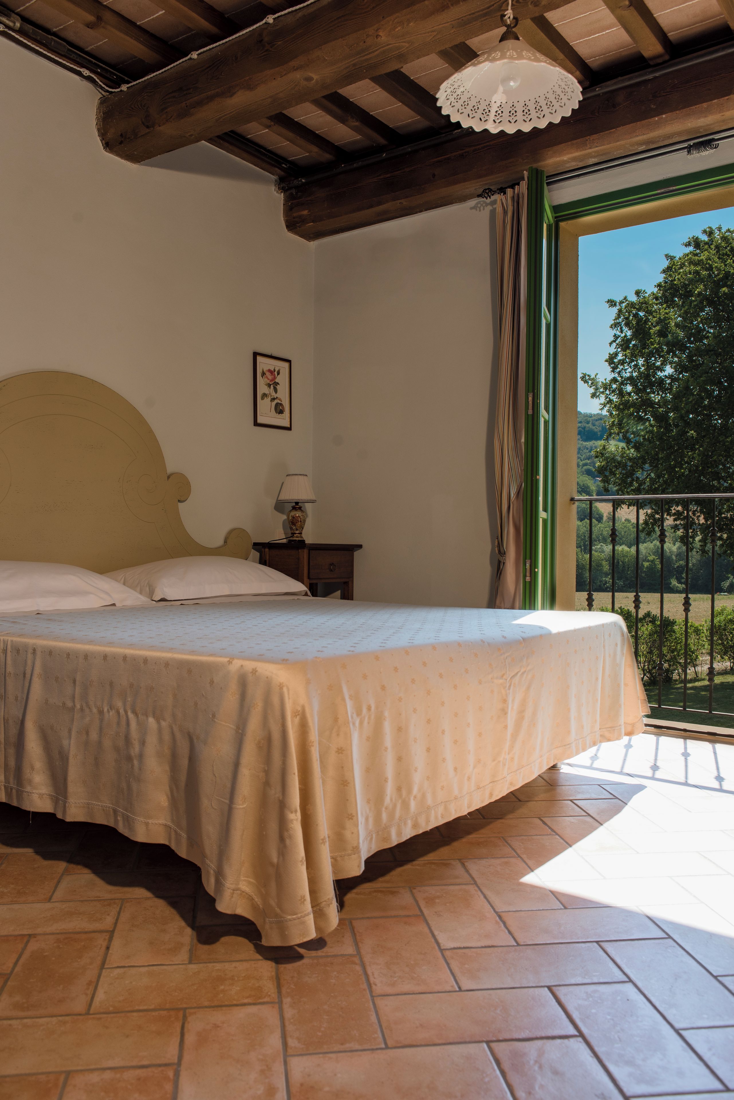 Un dormitorio con una cama, una mesita de noche, una lámpara, un cuadro enmarcado y vistas a la vegetación por una ventana.