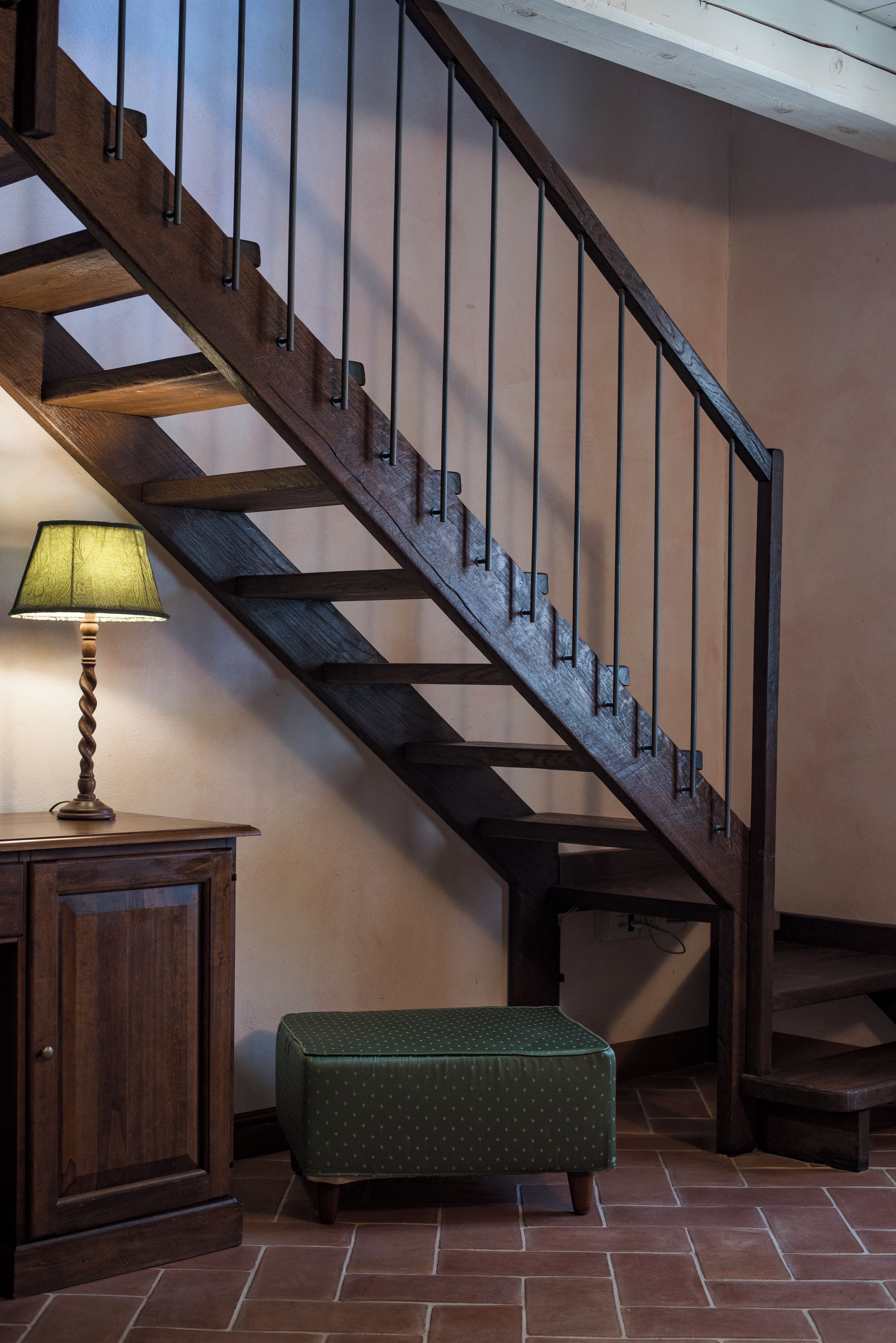 A wooden staircase with metal balusters, a green upholstered bench, and a lamp on a wooden cabinet.