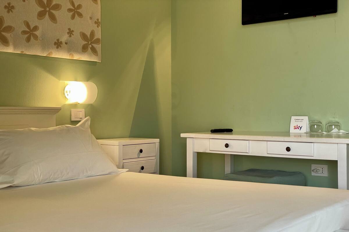 Una cama bien hecha con sábanas blancas, una mesita de noche con lámpara, un cajón pequeño y un cartel que indica una cámara con Sky WiFi.