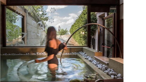 A person in a hot tub with a view of a garden and a building.