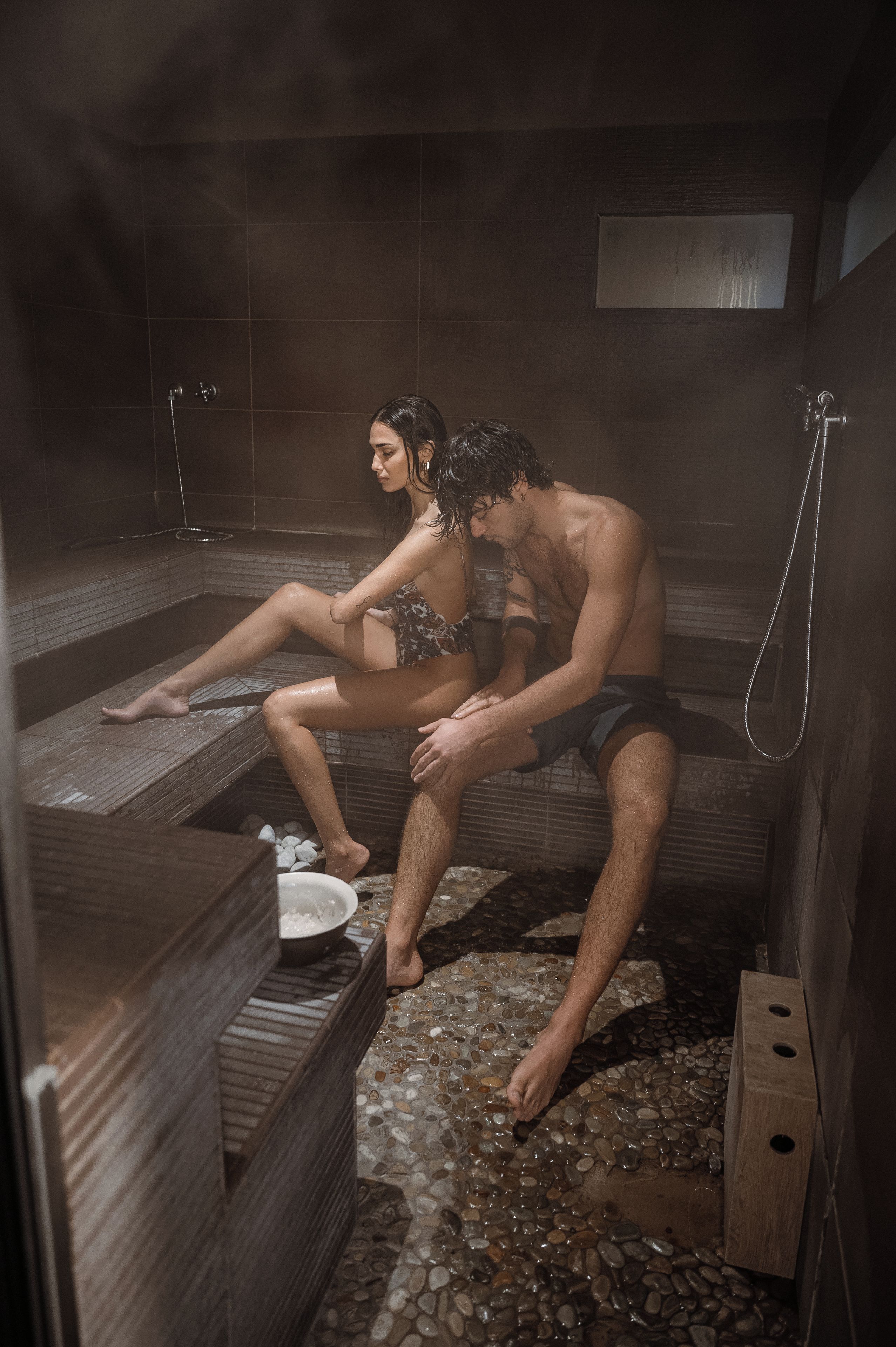 Uma sauna com duas pessoas sentadas em bancos, vapor visível e chão de azulejos.
