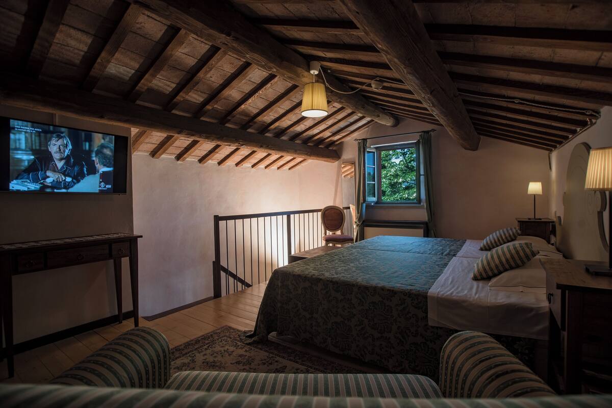 Un dormitorio acogedor con techo de madera, cama con ropa de cama a rayas, televisor en la pared y ventana con vista a árboles.