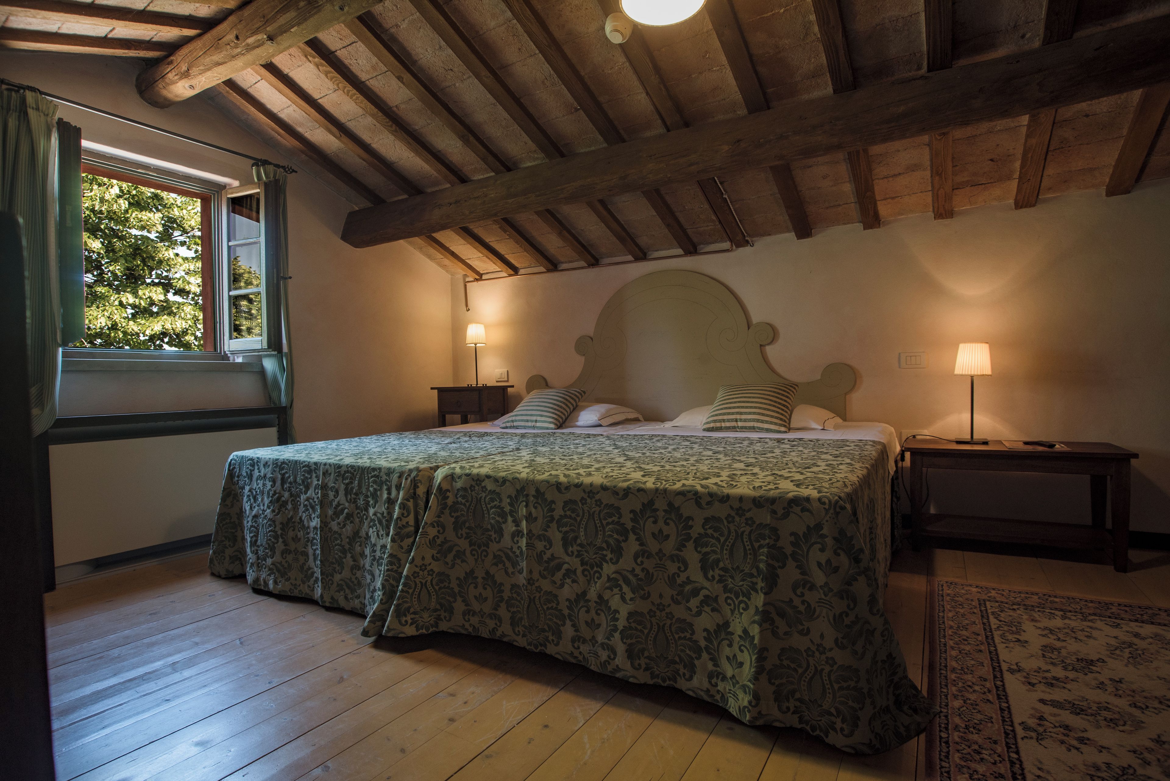 Ein Schlafzimmer mit Holzboden, einem Bett mit gemustertem Überwurf, zwei Nachttischlampen, einem Fenster mit Vorhängen und einer Holzzielung.