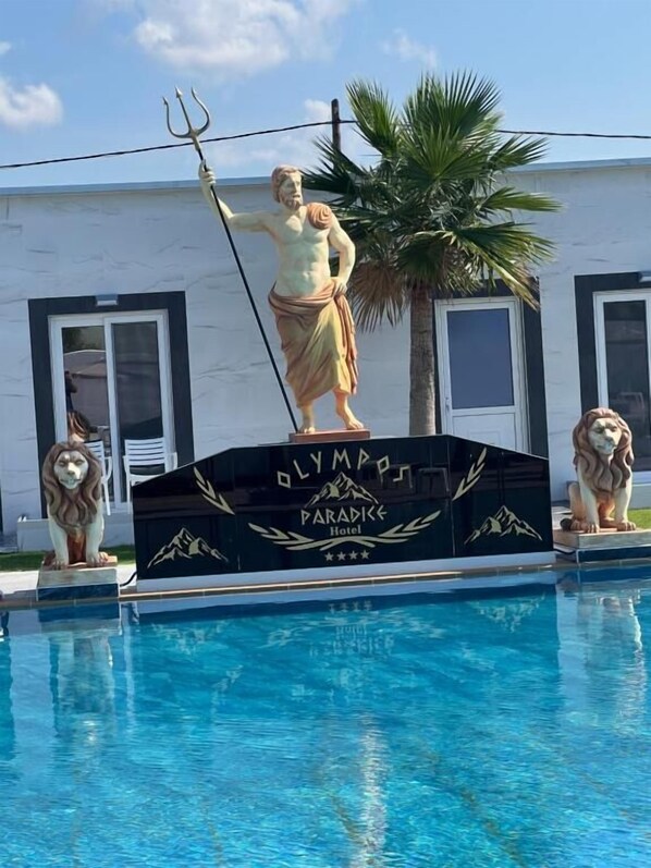 A statue of a figure holding a trident by a poolside, with two lion statues on either side. The background includes a building with the sign "Olympia Paradise Hotel". - Paradice Hotel Luxury Suites (Chania)