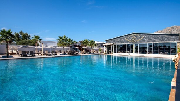 A clear blue pool with lounge chairs and umbrellas, a restaurant with a view, and palm trees in the background. - Paradice Hotel Luxury Suites (Chania)