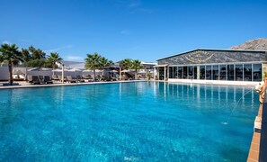 A clear blue pool with lounge chairs and umbrellas, a restaurant with a view, and palm trees in the background. - Paradice Hotel Luxury Suites (Chania)
