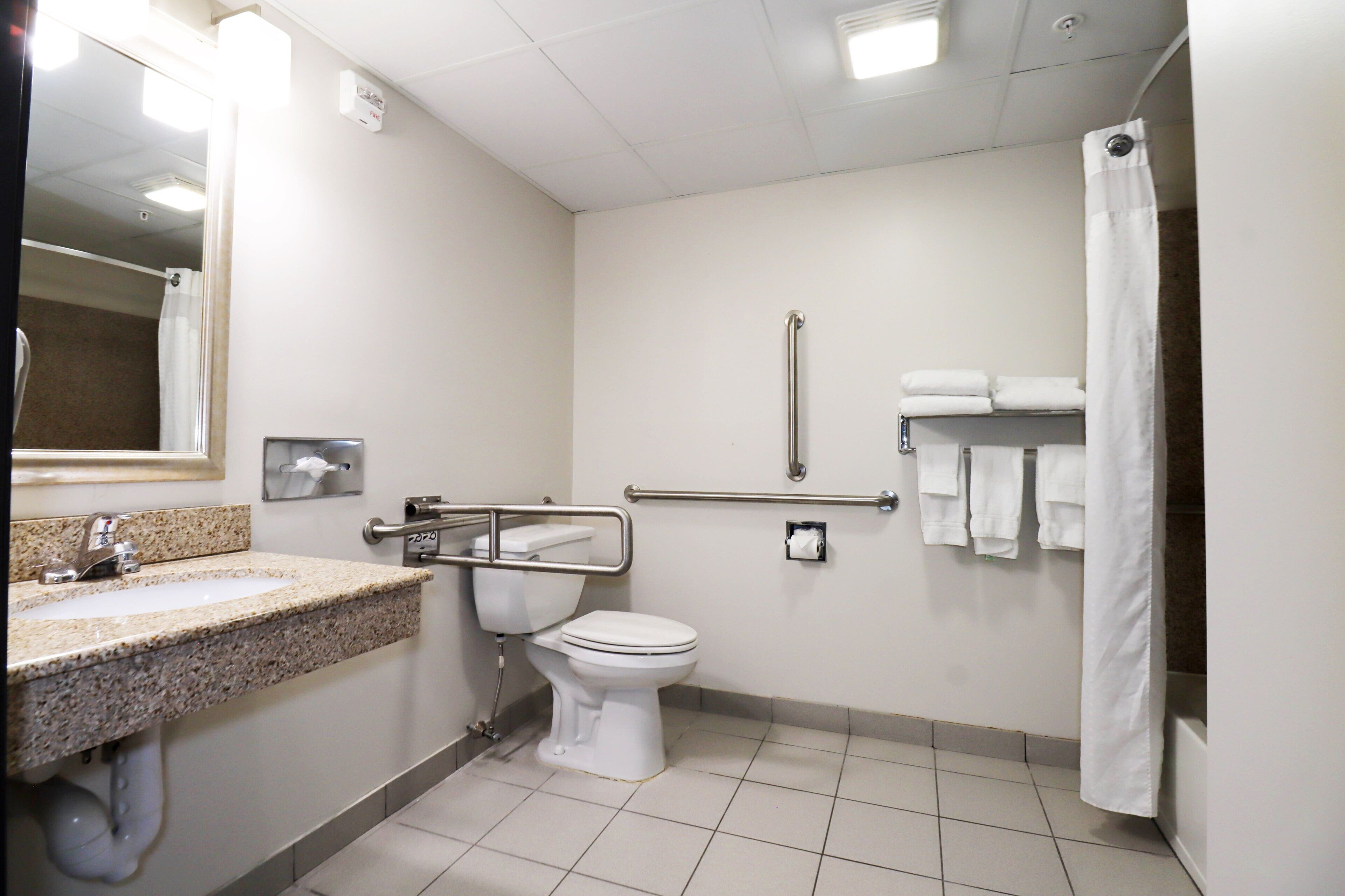 A standard hotel bathroom with a sink, toilet, shower, and handrails.