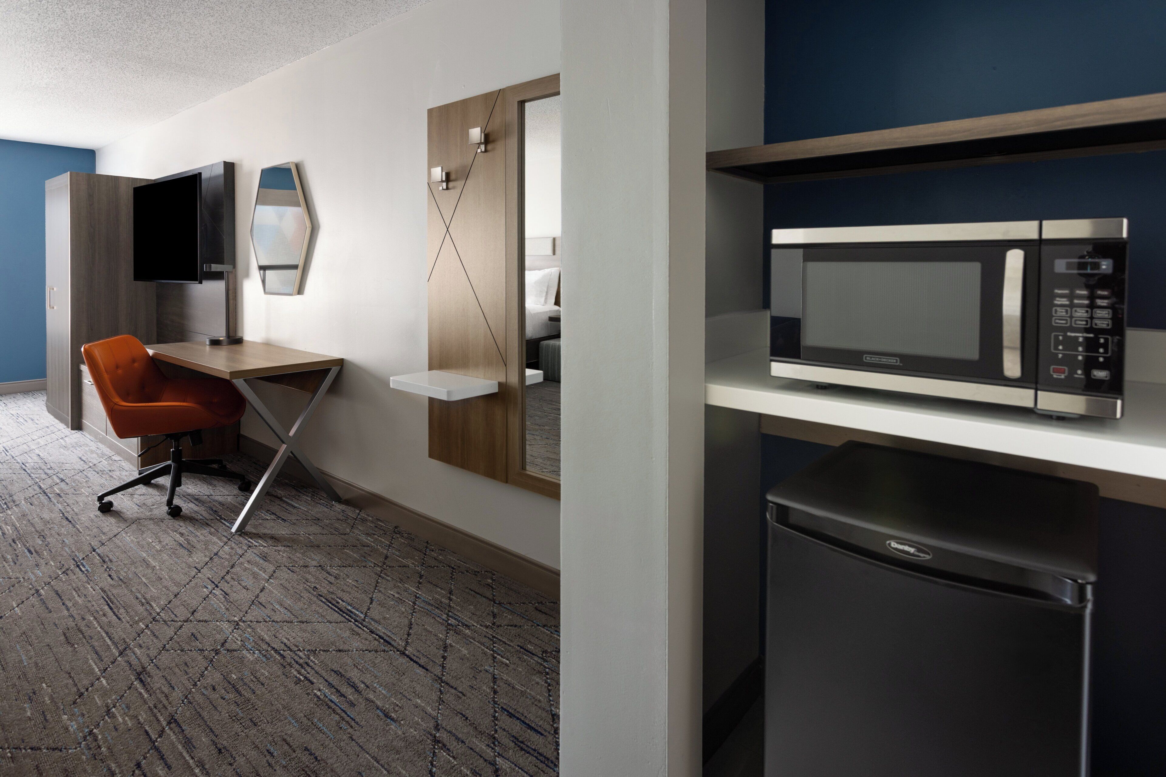 Una habitación de hotel moderna con escritorio, silla, televisor de pantalla plana, microondas y refrigerador.