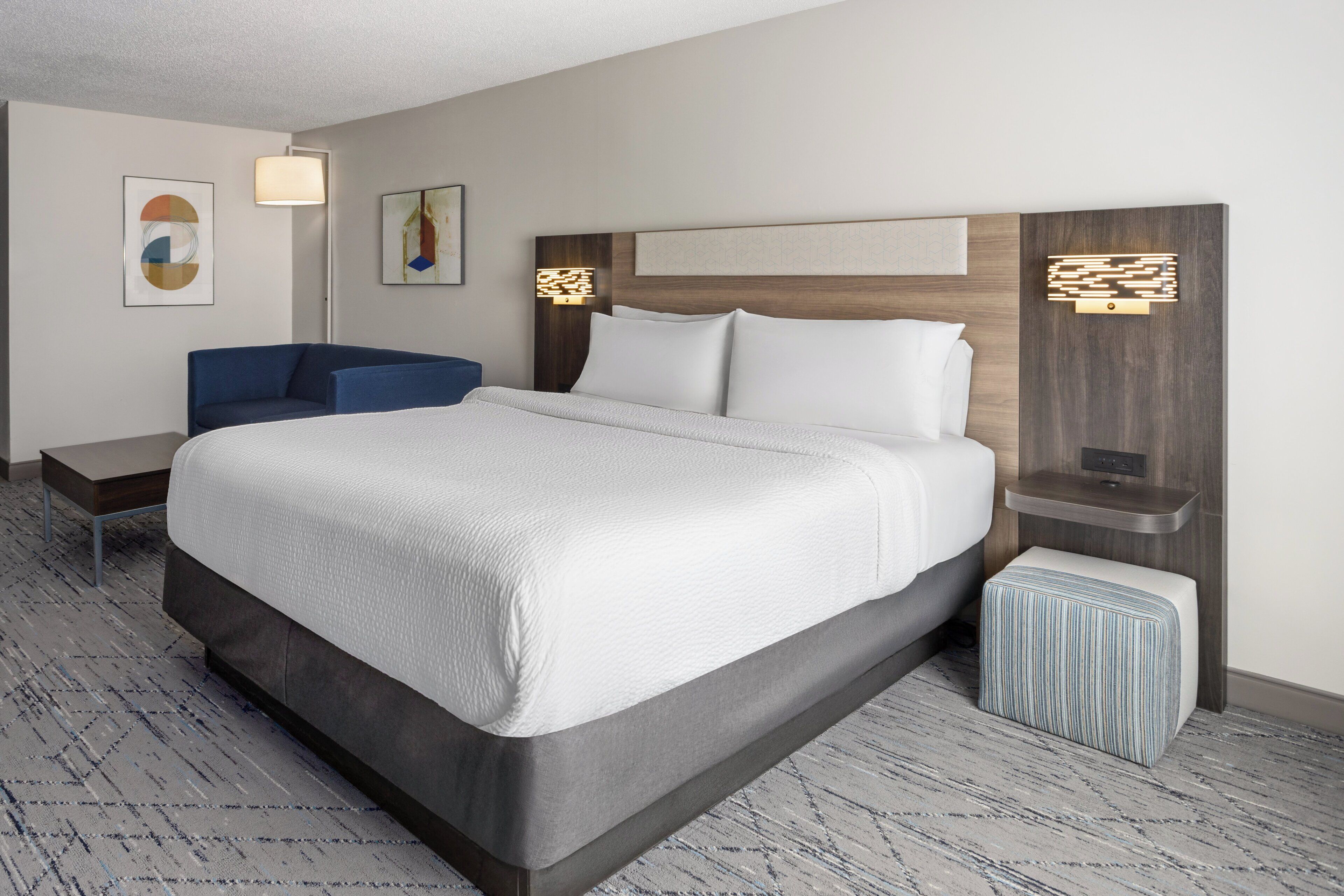 Ein Hotelzimmer mit einem großen Bett, einem blauen Sessel, einem Nachttisch und einem kleinen Hocker.