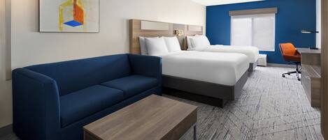Habitación de hotel con un sofá azul, una mesa de centro de madera, dos camas con ropa de cama blanca, una pared de acento azul y un ventanal grande.