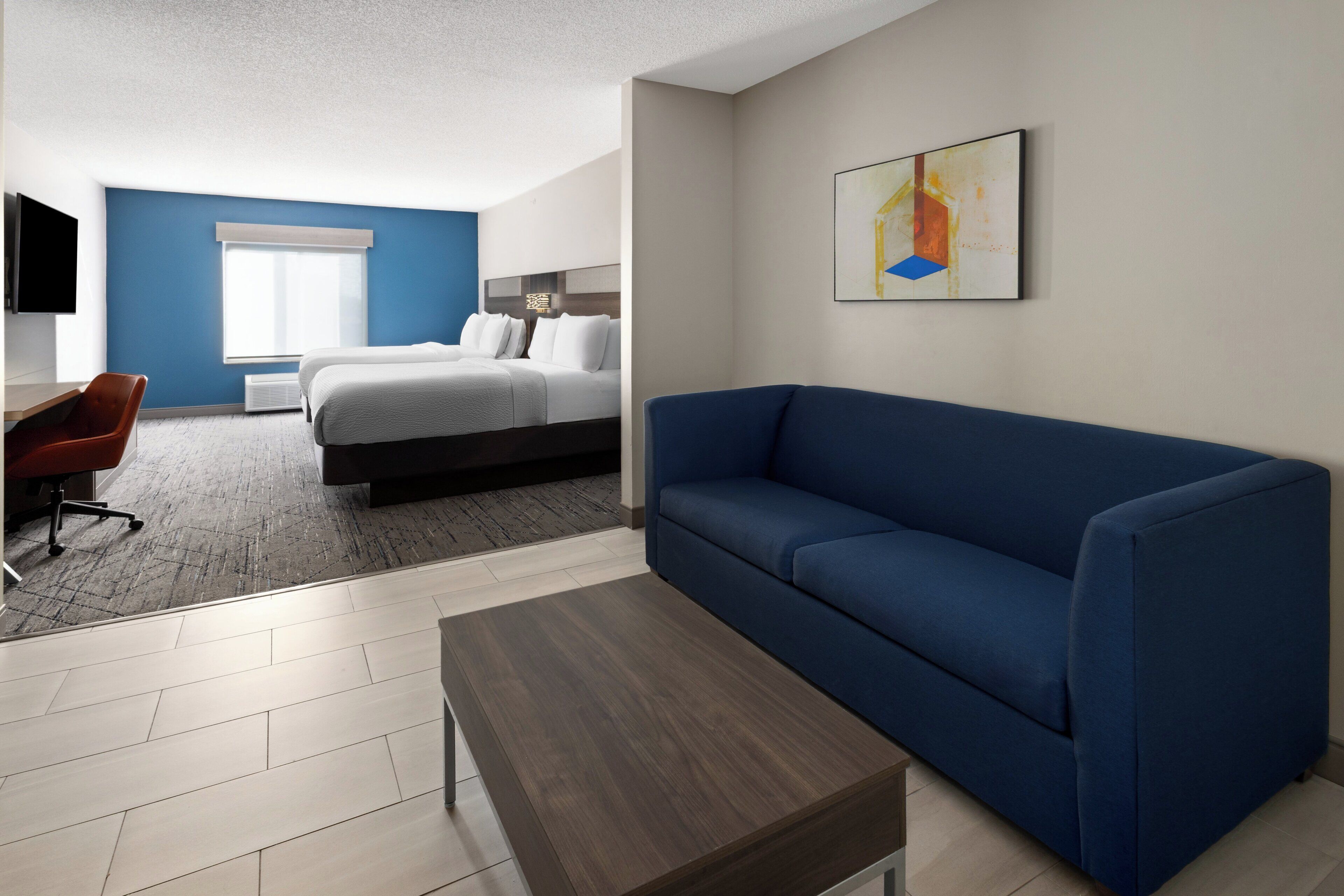 Ein modernes Hotelzimmer mit einem blauen Sofa, einem Holztisch, einem Schreibtisch mit Stuhl, einem Bett mit weißer Bettwäsche und einem großen Fenster mit Jalousien.