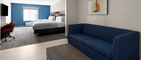 A modern hotel room with a blue sofa, a wooden coffee table, a desk with a chair, a bed with white linens, and a large window with blinds.