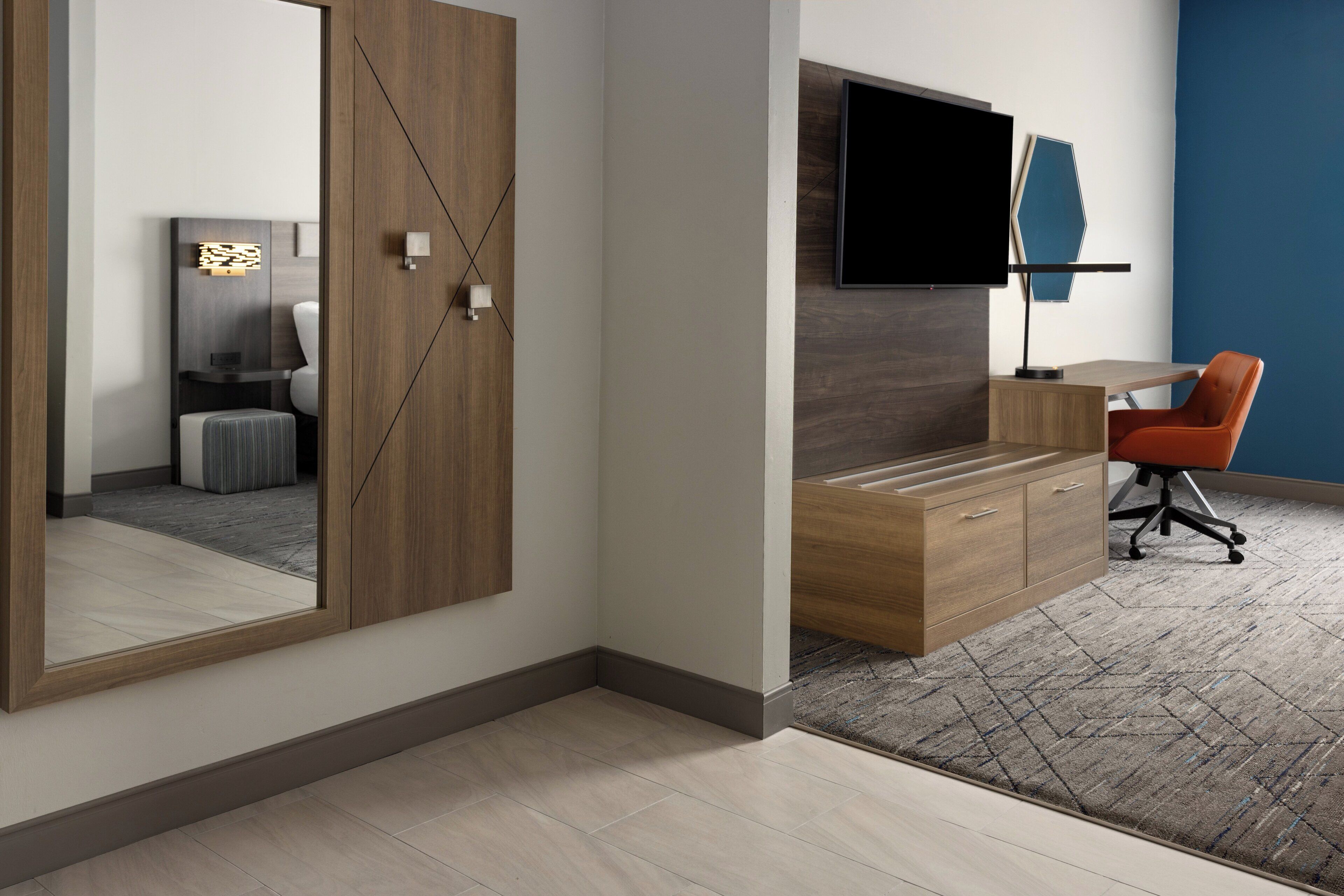 A modern bedroom with a wooden wardrobe, a desk with a chair, a flat-screen TV, and a large mirror.