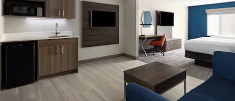 Una habitación de hotel moderna con kitchenette, un televisor de pantalla plana montado en la pared, un escritorio con una silla, una cama con cabecera azul y un sofá azul.