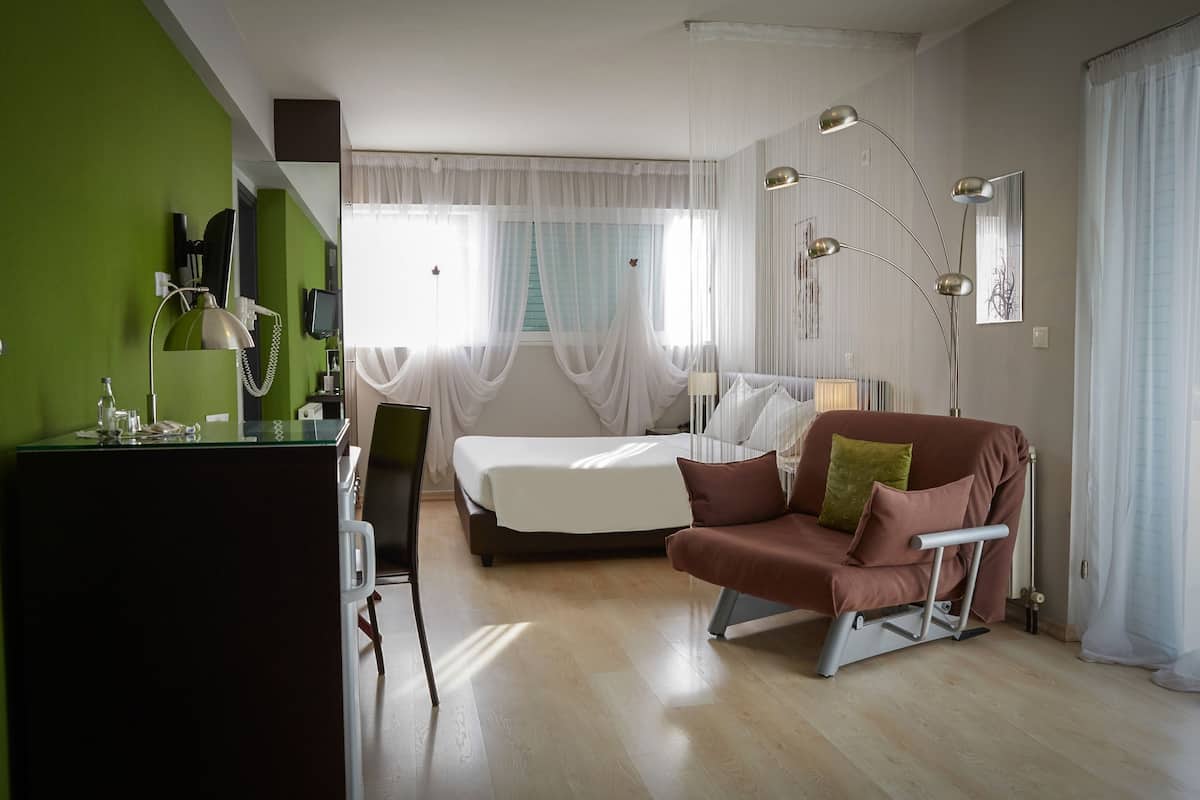Une chambre d’hôtel moderne, équipée d’un lit, d’un canapé, d’un bureau avec une lampe et d’une grande fenêtre agrémentée de rideaux légers.
